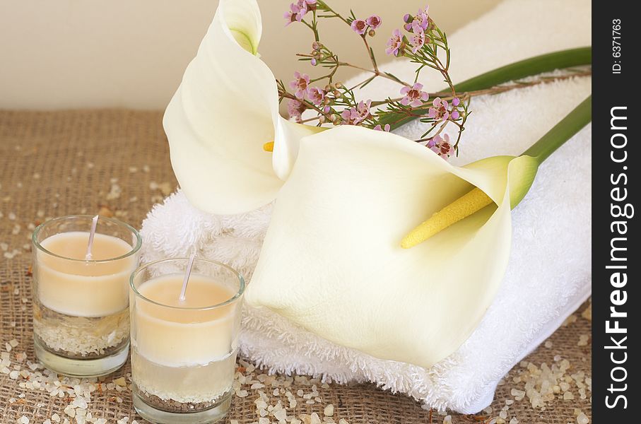 Relaxing spa scene with a white rolled up towel, white lillies, small pink flowers, beautiful handmade candles and bath salts