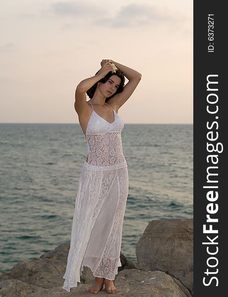 Young woman wearing white dress during sunset at beach staying on the rock. Young woman wearing white dress during sunset at beach staying on the rock