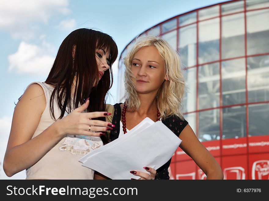 Two young women having a discussion, modern office building on the background.