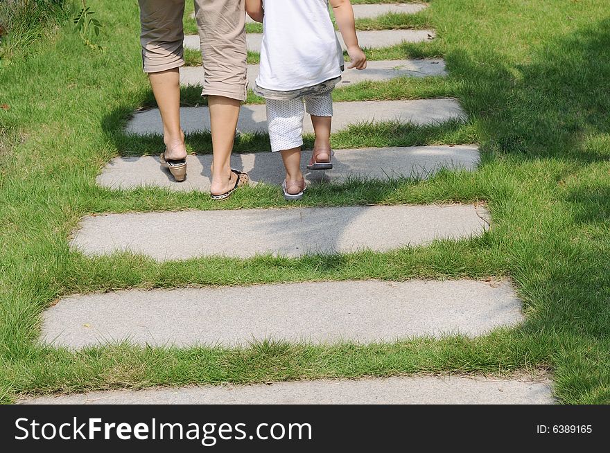 A toddler and his granny walk on the stone paved footpath in the meadow in a park.