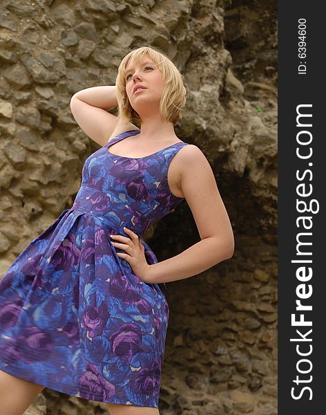 Stylish blond girl in purple dress posing beside ancient castle wall