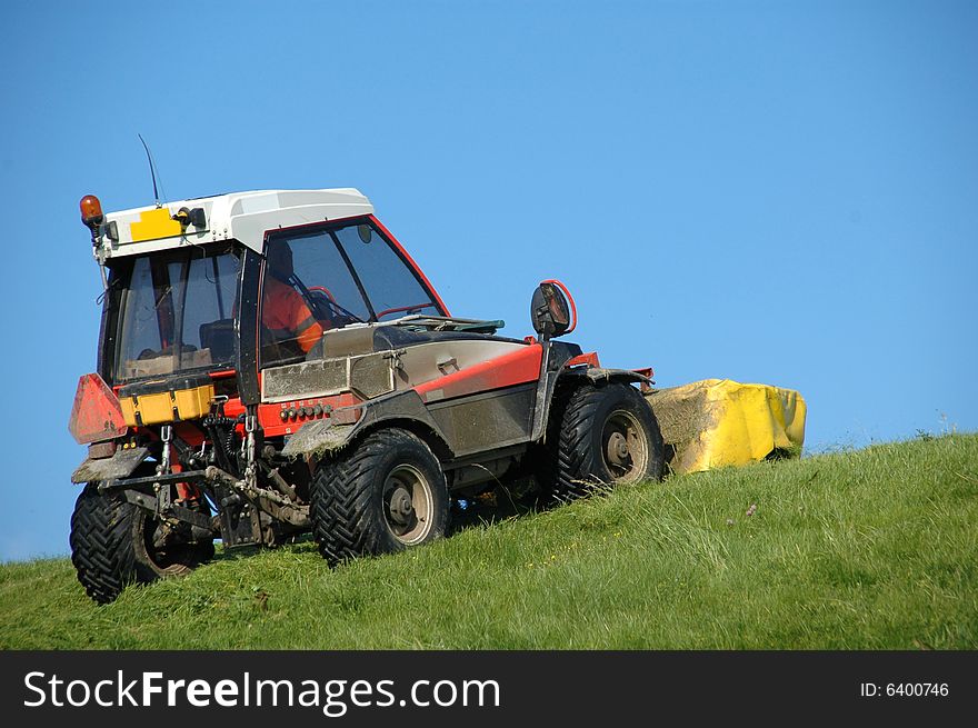 Big lawnmower mowing the grass on top of a dike against a clear blue sky. Big lawnmower mowing the grass on top of a dike against a clear blue sky