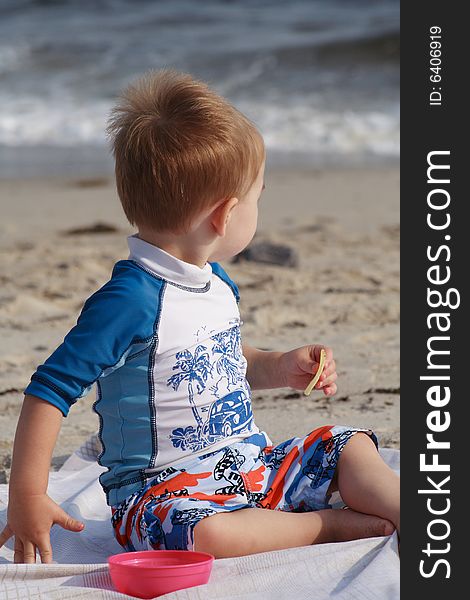 A toddler boy at the beach sitting on a beach blanket eating a snack. A toddler boy at the beach sitting on a beach blanket eating a snack.