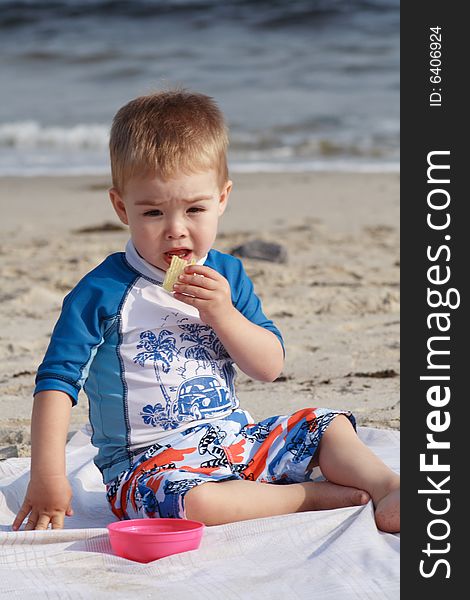 A toddler boy at the beach sitting on a beach blanket eating a snack. A toddler boy at the beach sitting on a beach blanket eating a snack.