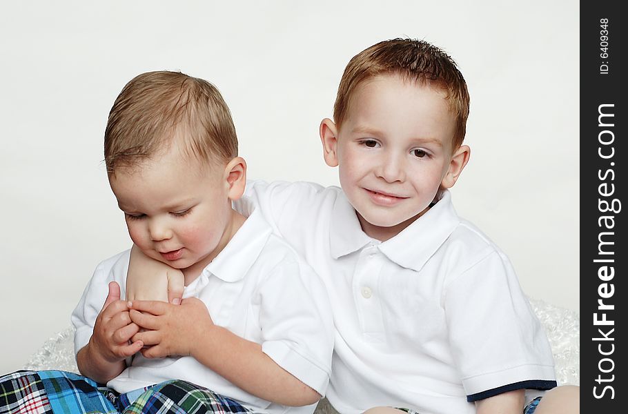 Two brothers are posing for a picture in a studio.  They are wearing matching outfits and the older boy is smiling at the camera with his arm around his younger brother.  The younger boy is looking down at his hands.  Horizontally framed shot. Two brothers are posing for a picture in a studio.  They are wearing matching outfits and the older boy is smiling at the camera with his arm around his younger brother.  The younger boy is looking down at his hands.  Horizontally framed shot.