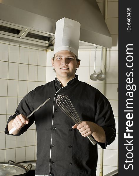 Smiling chef standing in a kitchen holding a knife and a whisk. Vertically framed photo. Smiling chef standing in a kitchen holding a knife and a whisk. Vertically framed photo.