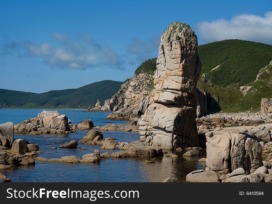 A small bay with rocks and many stones. On background are green cape and blue sky. Japanese Sea. Summer, sunny day. A small bay with rocks and many stones. On background are green cape and blue sky. Japanese Sea. Summer, sunny day.