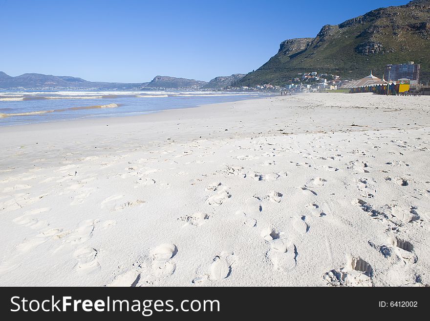 A beautiful white sandy beach with footprints. The seashore, some mountains and colourful dressing huts are visible in the background.