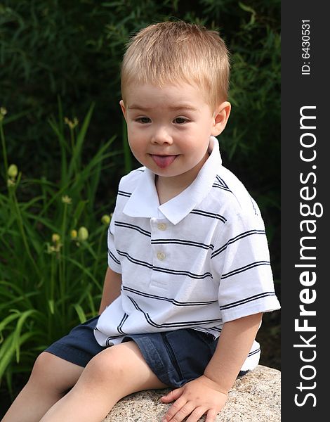A toddler boy wearing a striped golf shirt and shorts sitting on a rock and showing his tongue. A toddler boy wearing a striped golf shirt and shorts sitting on a rock and showing his tongue.