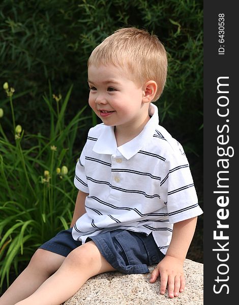 A Toddler boy wearing a striped white golf shirt sitting on a rock looking away from the camera. A Toddler boy wearing a striped white golf shirt sitting on a rock looking away from the camera.