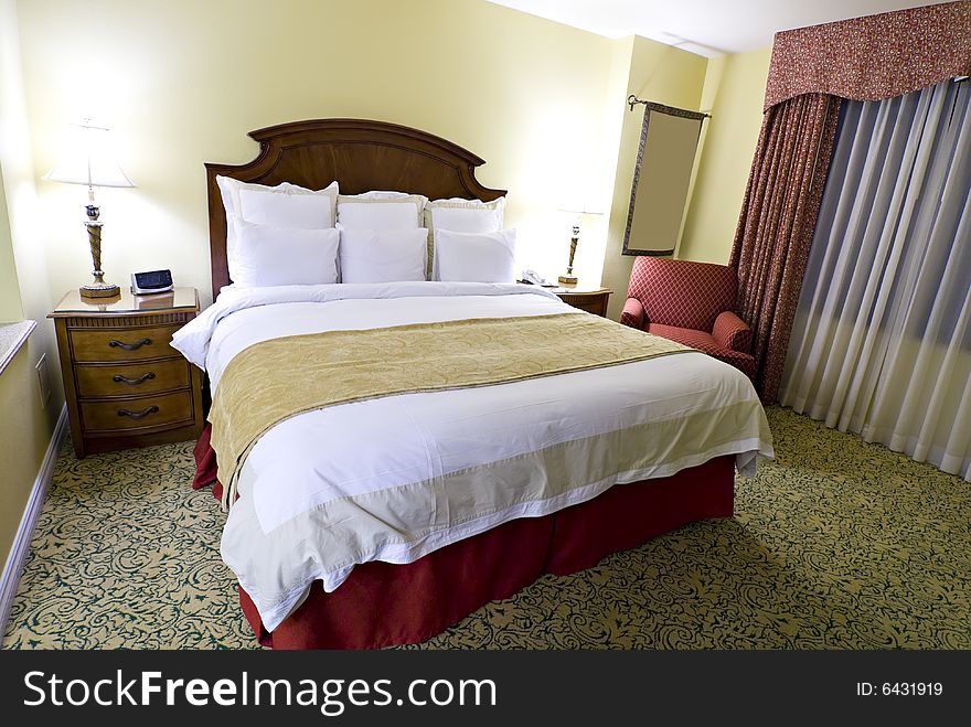 View of large king sized bed with nightstand and lamp and curtained window in background. View of large king sized bed with nightstand and lamp and curtained window in background