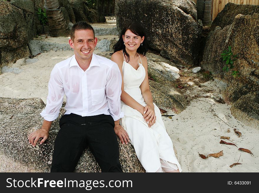 Happy bride and groom sitting on a rock at the beach.