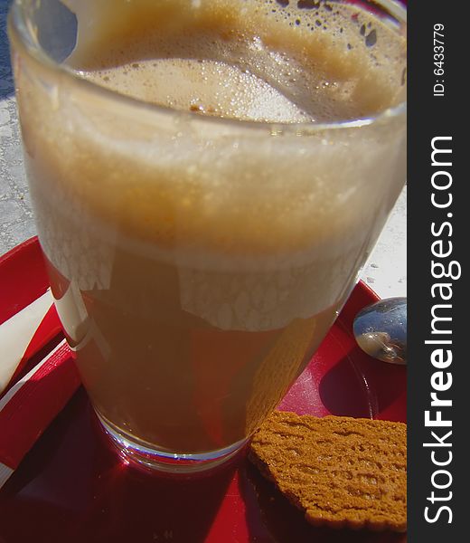 Close-up photo of the transparent cappuccino cup on red plate, with biscuit and spoon, on the table; view from above. Close-up photo of the transparent cappuccino cup on red plate, with biscuit and spoon, on the table; view from above
