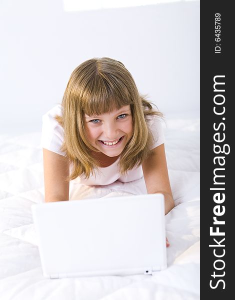 Young smiling girl lying on a bed and does something on laptop. She's looking at camera. Young smiling girl lying on a bed and does something on laptop. She's looking at camera.