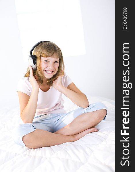 Young smiling girl sitting cross-legged on a bed and listening music by earphones. She's looking at camera. Young smiling girl sitting cross-legged on a bed and listening music by earphones. She's looking at camera.