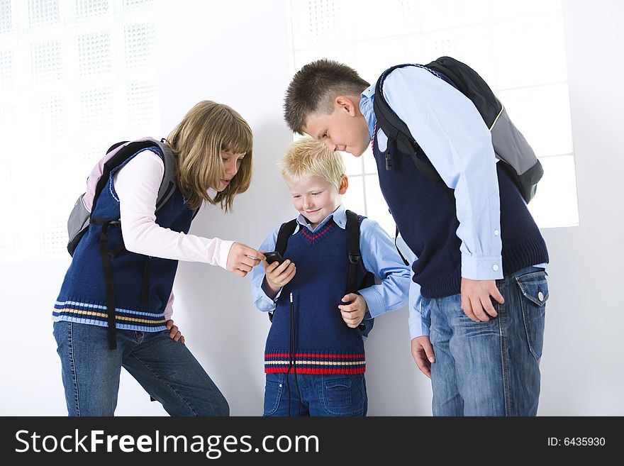 Three schoolchildren standing at corridor in front of window. Younger schoolchild showing something in cellphone. Three schoolchildren standing at corridor in front of window. Younger schoolchild showing something in cellphone.