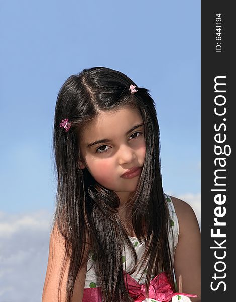 Face of a young little girl with dark hair looking serious. Set against a blue sky with clouds. Face of a young little girl with dark hair looking serious. Set against a blue sky with clouds.