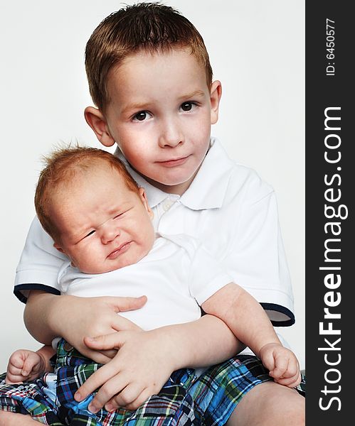 An older and a younger brother are posing together for a picture in a studio.  The older boy is smiling at the camera, and the infant is staring at the camera.  Vertically framed shot. An older and a younger brother are posing together for a picture in a studio.  The older boy is smiling at the camera, and the infant is staring at the camera.  Vertically framed shot.