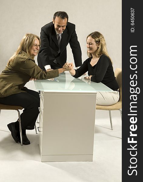 Two women arm wrestling across a desk while a man stands over watching. They are all smiling. Vertically framed photo.