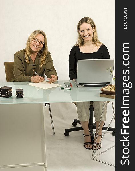 Two people are sitting together in a room at a table. The two woman are looking and smiling at the camera. It appears to be a business meeting and there is a laptop on the table in front of the younger woman. Vertically framed shot. Two people are sitting together in a room at a table. The two woman are looking and smiling at the camera. It appears to be a business meeting and there is a laptop on the table in front of the younger woman. Vertically framed shot.