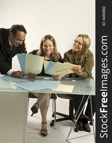 Three people are together in a room at a table. It appears to be a business meeting. They are all looking down at pieces of paper in their hands and smiling. Vertically framed shot. Three people are together in a room at a table. It appears to be a business meeting. They are all looking down at pieces of paper in their hands and smiling. Vertically framed shot.