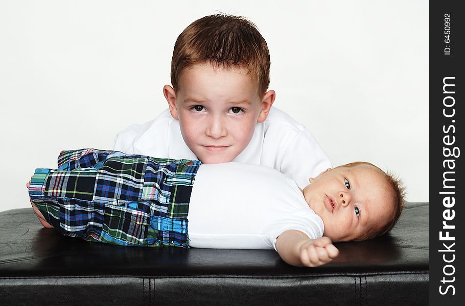 An older and a younger brother are posing together for a picture in a studio.  The older boy is smiling at the camera, and the infant is staring at the camera.  Horizontally framed shot. An older and a younger brother are posing together for a picture in a studio.  The older boy is smiling at the camera, and the infant is staring at the camera.  Horizontally framed shot.
