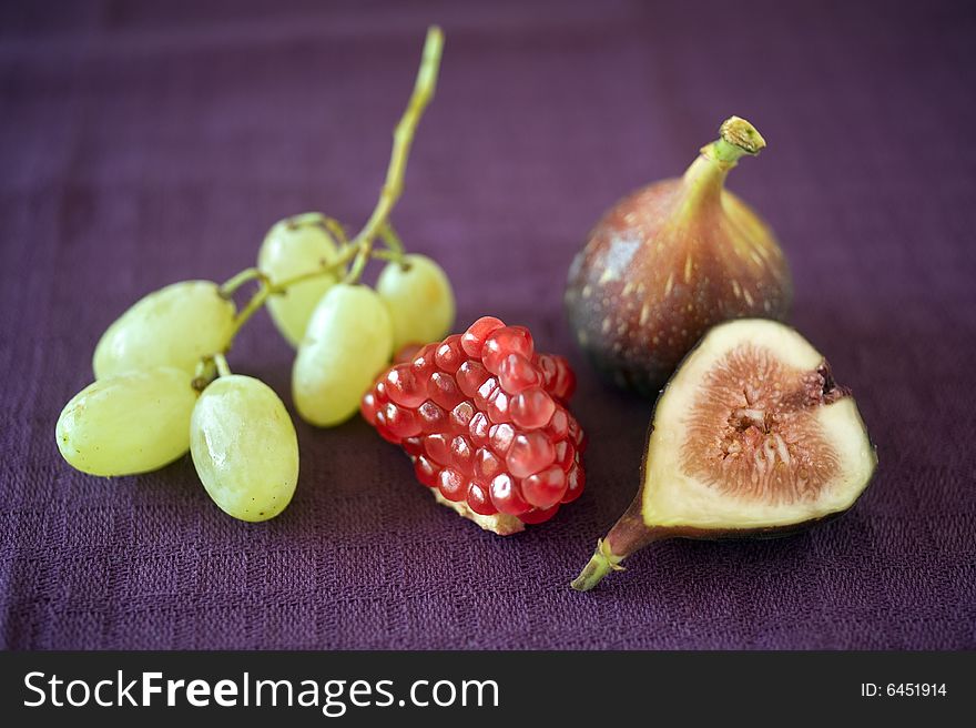 Still life image of a part of pomegranate, grapes, a whole fig and a slice of fig on purple cloth. Still life image of a part of pomegranate, grapes, a whole fig and a slice of fig on purple cloth
