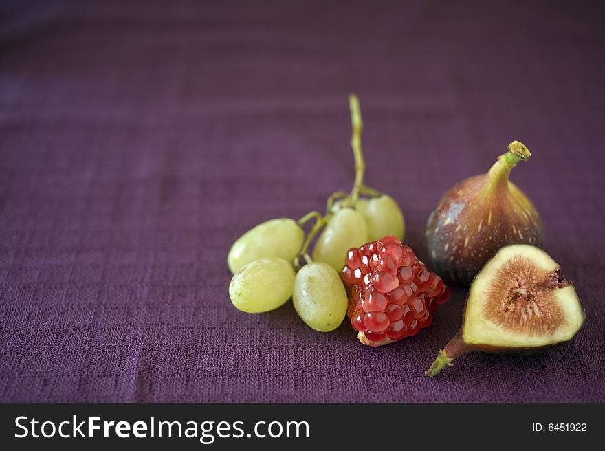 Still life image of a part of pomegranate, grapes, a whole fig and a slice of fig on purple cloth. Still life image of a part of pomegranate, grapes, a whole fig and a slice of fig on purple cloth