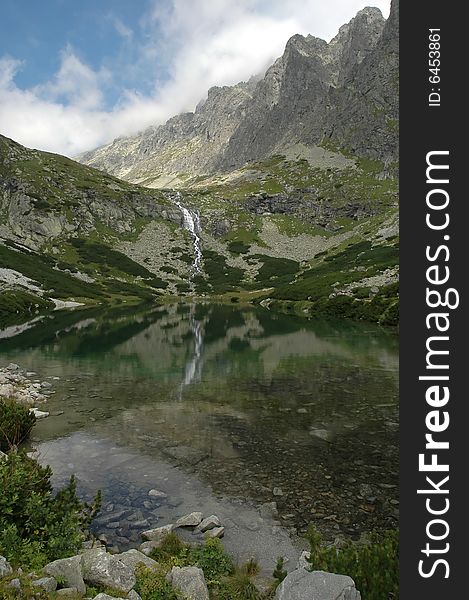 Cold waterfall feeding crystal clear lake under sharp mountains at the end of summer. Cold waterfall feeding crystal clear lake under sharp mountains at the end of summer.