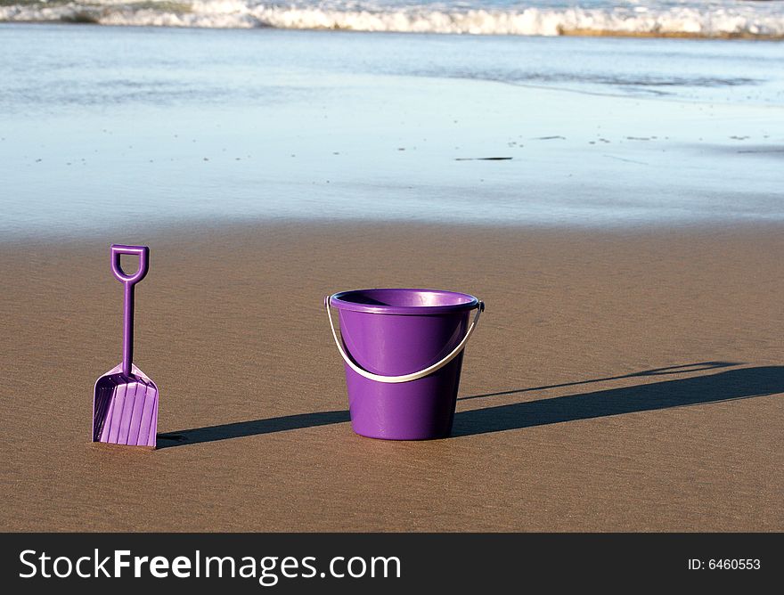 A purple plastic bucket and spade on the beach with the ocean behind them coming in. A purple plastic bucket and spade on the beach with the ocean behind them coming in
