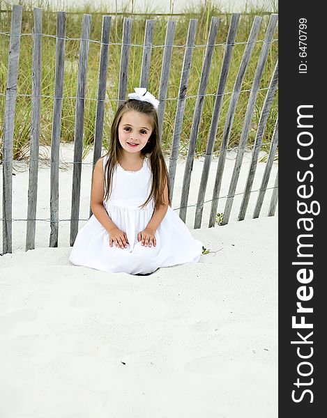 Beautiful young brunettte girl sitting on the sand dressed in a white dress by a wooden fence. Beautiful young brunettte girl sitting on the sand dressed in a white dress by a wooden fence.