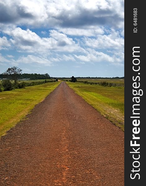 Long, empty country road in the middle of nowhere with grass fields on either side and cloudy blue sky