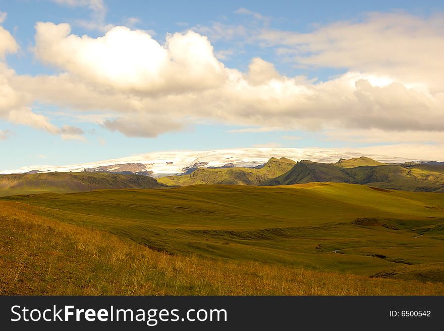 Glacier with a grassland in the foreground. Cloudy blue sky.