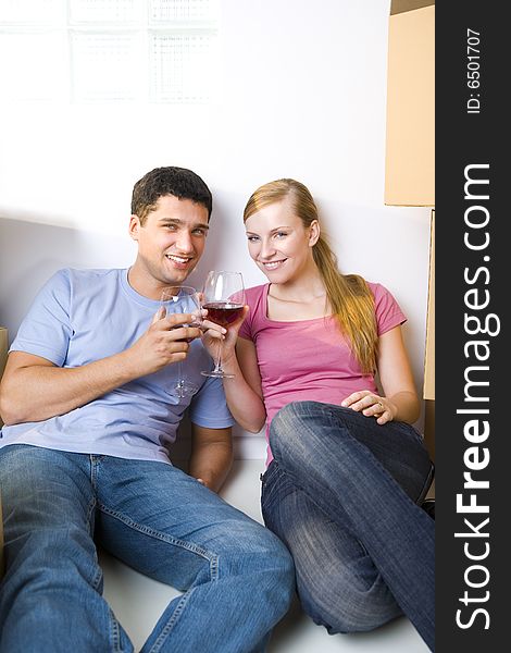 Young couple sitting between cardboard boxes and drinking wine. They're smiling and looking at camera. Front view. Young couple sitting between cardboard boxes and drinking wine. They're smiling and looking at camera. Front view.