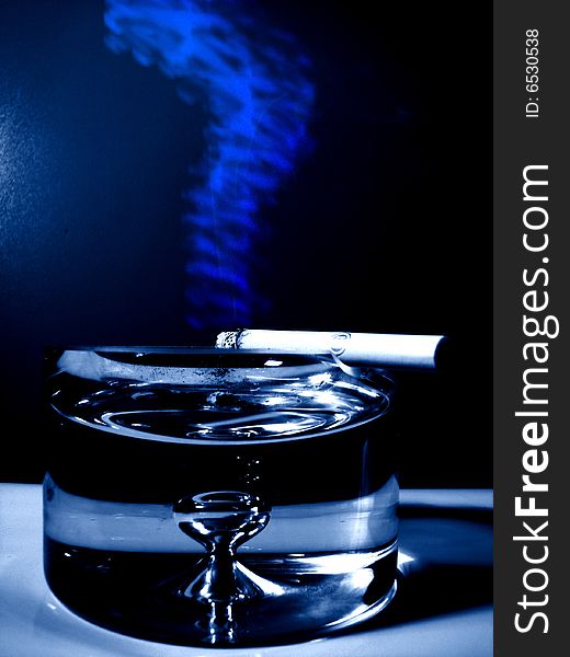 Cigarette with blue smoke on a big glass ash-tray in navy blue color. Cigarette with blue smoke on a big glass ash-tray in navy blue color.