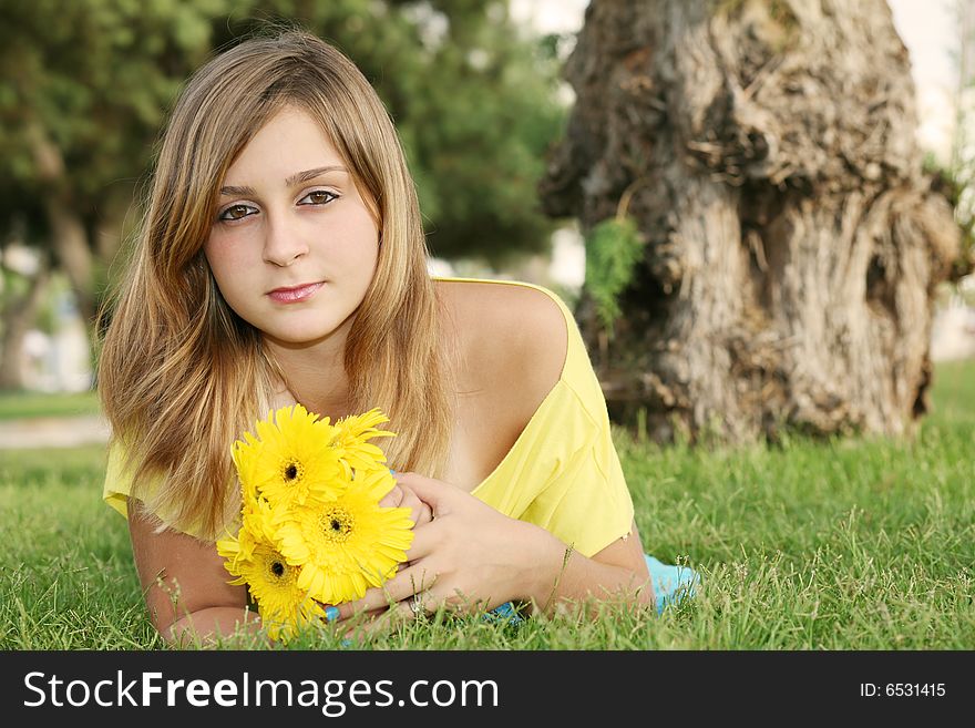 A young beautiful girl rests with the bouquet of yellow flowers on a green grass in a city park