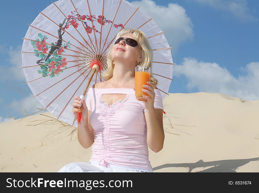 A young blonde woman sits on sand with glass of juice and umbrella. A young blonde woman sits on sand with glass of juice and umbrella