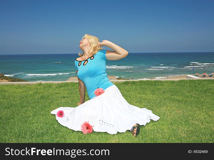 A young beautiful blonde sits on a green grass on a background blue sky and dark blue sea. A young beautiful blonde sits on a green grass on a background blue sky and dark blue sea
