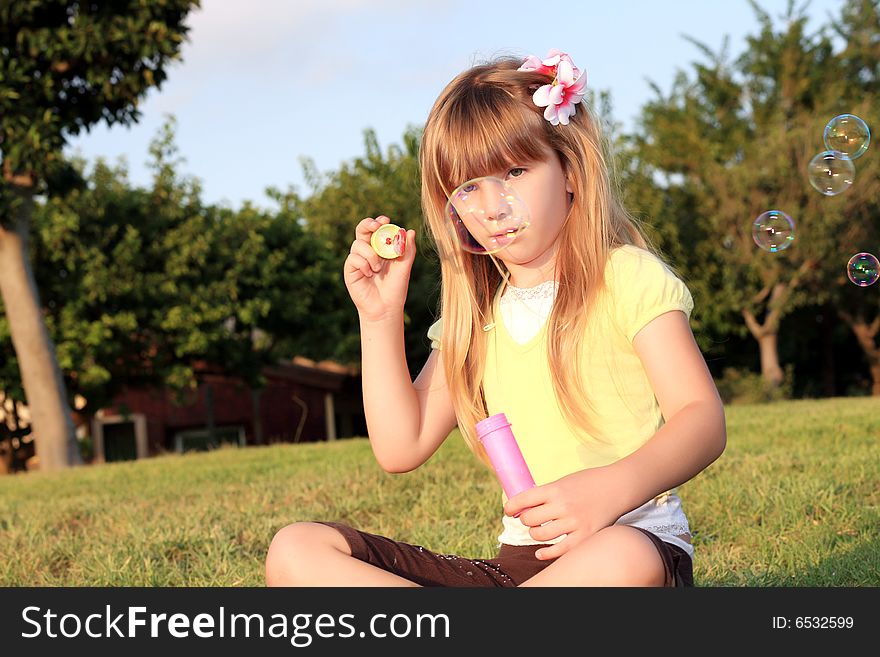 A little beautiful blonde sits on a green grass on a background blue sky and allows soap-bubbles. A little beautiful blonde sits on a green grass on a background blue sky and allows soap-bubbles