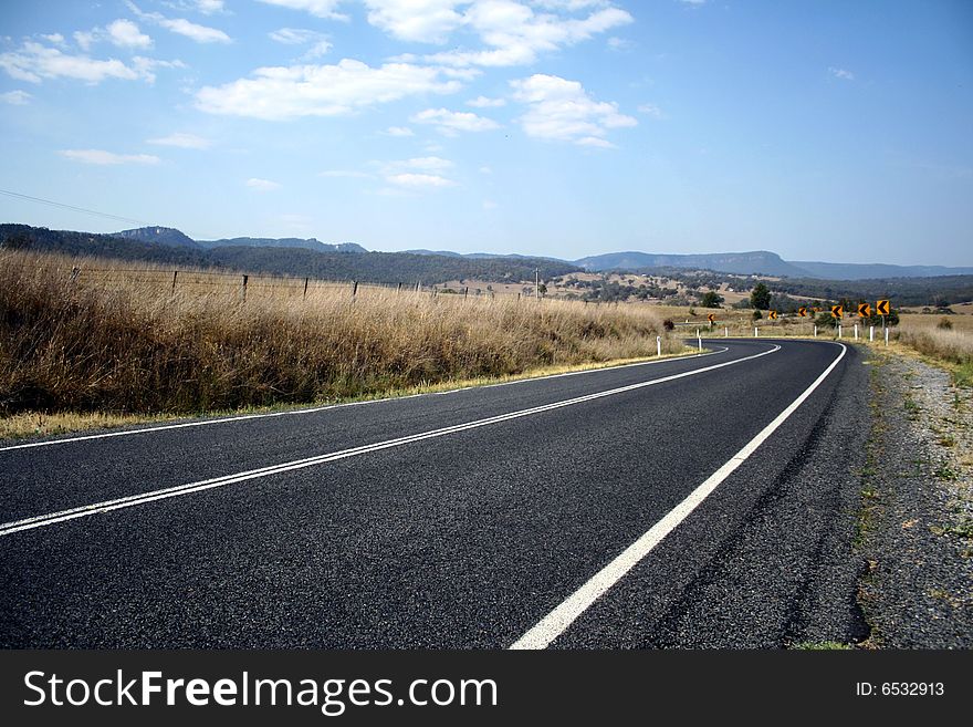 A long tar road meandering to the left, surrounded by a landscape of dry shrub. A long tar road meandering to the left, surrounded by a landscape of dry shrub.