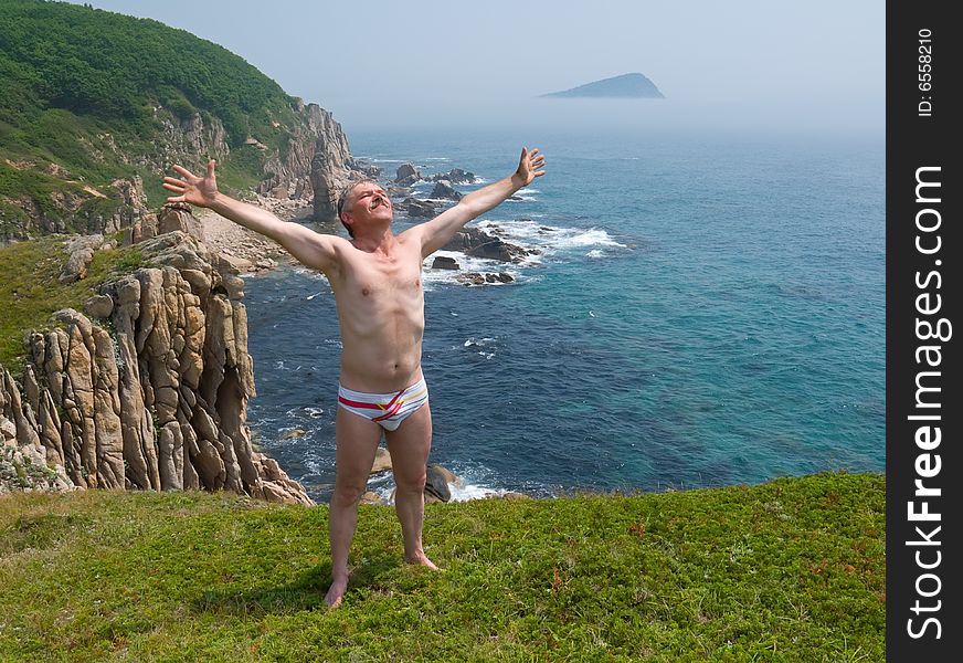 A  man sunburns on flat green top of cliff on seacoast. On background is small bay with rocks and stones and small island in fog. A  man sunburns on flat green top of cliff on seacoast. On background is small bay with rocks and stones and small island in fog.