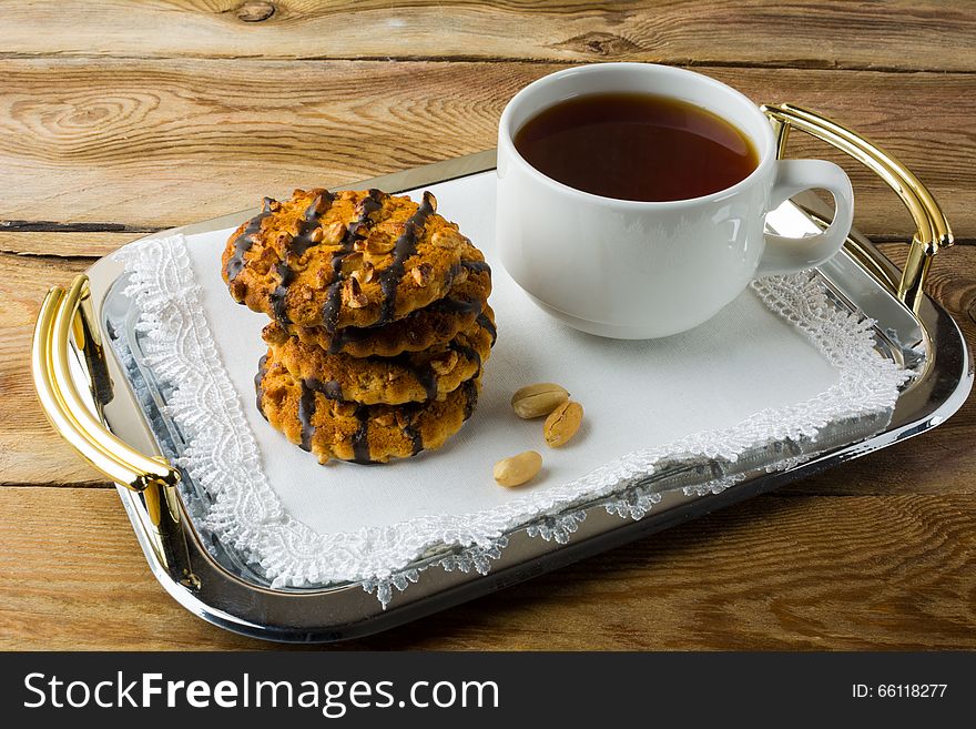 Chocolate icing cookies with peanuts and cup of tea on a metal serving tray with handles covered with a white lace napkin. Breakfast biscuits and tea.