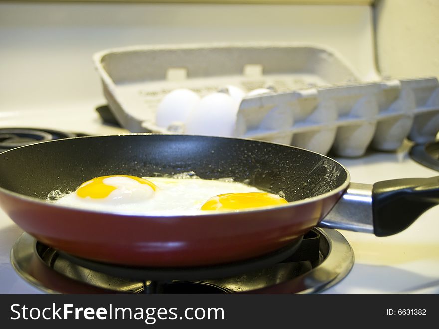 Two eggs frying in a small skillet with a carton of eggs in the background. Two eggs frying in a small skillet with a carton of eggs in the background.