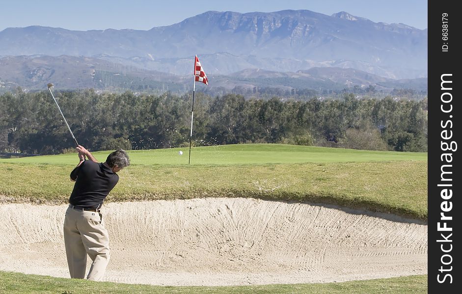 Golfer hitting out of bunker, ball in flight to pin, blue sky and mountains in background. Golfer hitting out of bunker, ball in flight to pin, blue sky and mountains in background