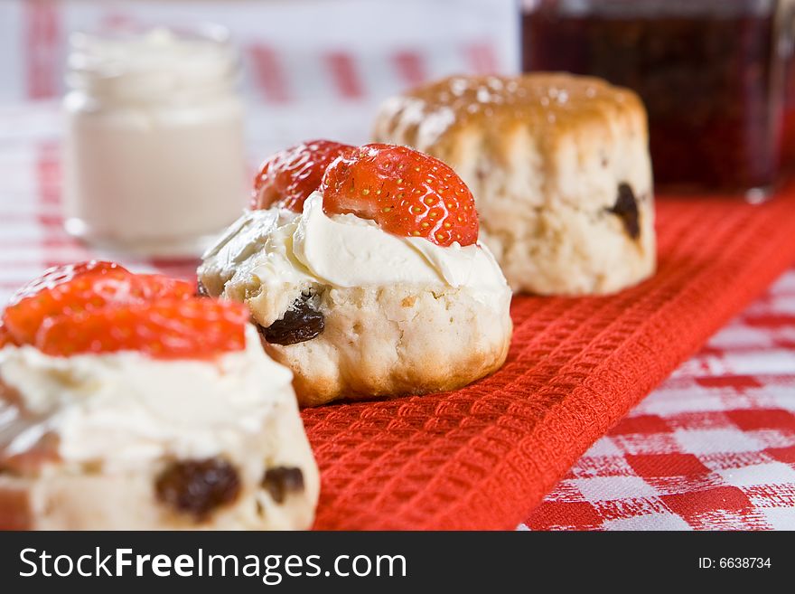 A line of scone halves with strawberries, clotted cream and a jar of raspberry jam on a red cloth. A line of scone halves with strawberries, clotted cream and a jar of raspberry jam on a red cloth