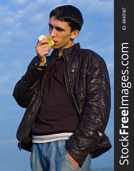 Young man eats an apple against the blue and cloudy sky, thinking about something. Young man eats an apple against the blue and cloudy sky, thinking about something