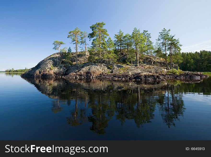 A small stony island rises from blue water with some pine trees on its surface