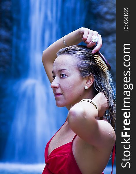 Young blond woman brushing her hair, long exposed cascade as background. Young blond woman brushing her hair, long exposed cascade as background