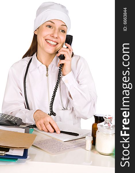 The smiling young woman-doctor speaks by phone near a table with records and medicines. The smiling young woman-doctor speaks by phone near a table with records and medicines