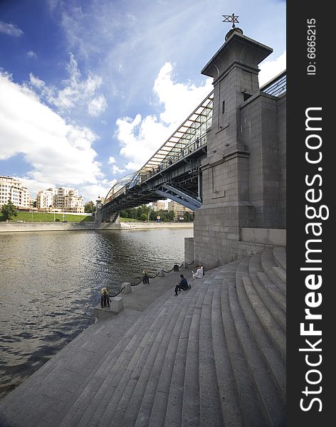 Grey bridge over the river and some people sitting on the bank of this river and clouds in the sky